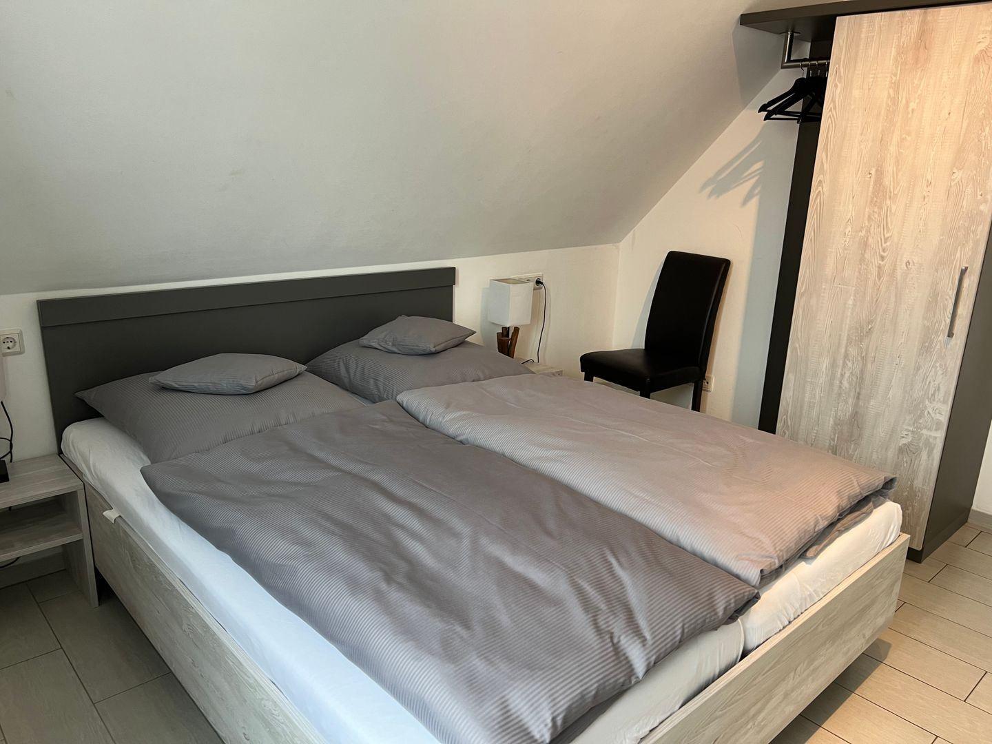 Doppelbett mit grauer Bettwäsche, Schrank und Stuhl in einem Schlafzimmer mit geneigtem Dach.