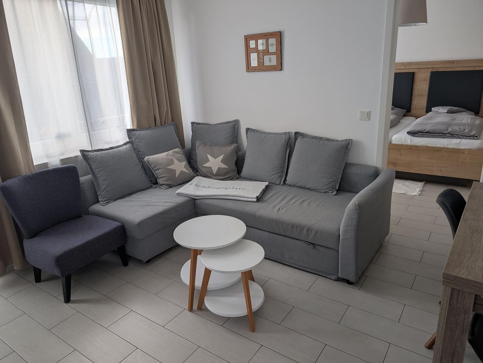 Ein Wohnzimmer mit grauem Sofa, Sitzgelegenheit und Blick in das Schlafzimmer.