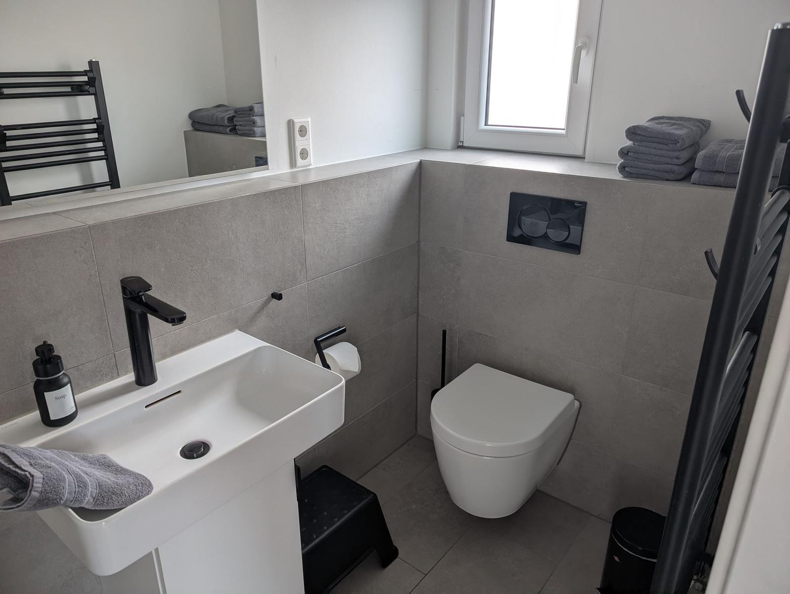 Badezimmer mit Waschbecken, Toilette und Fenster. Graue Fliesen und schwarze Accessoires.