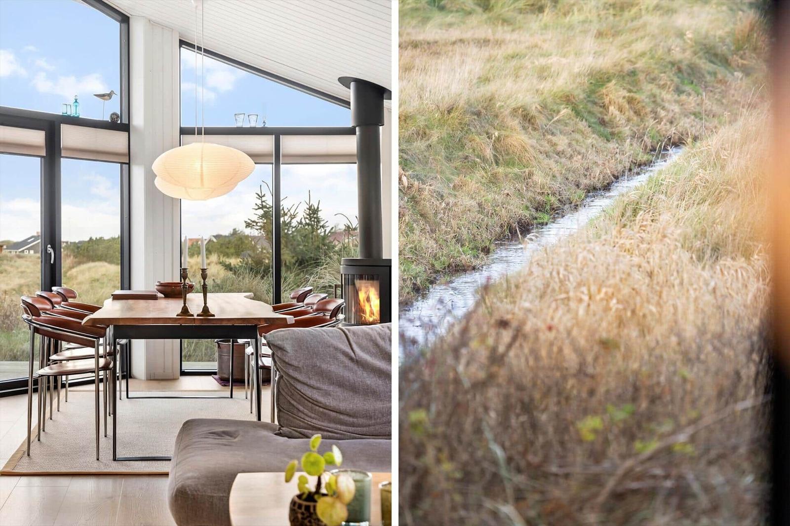 Modern dining room with large windows and fireplace. Outside view shows a small stream.