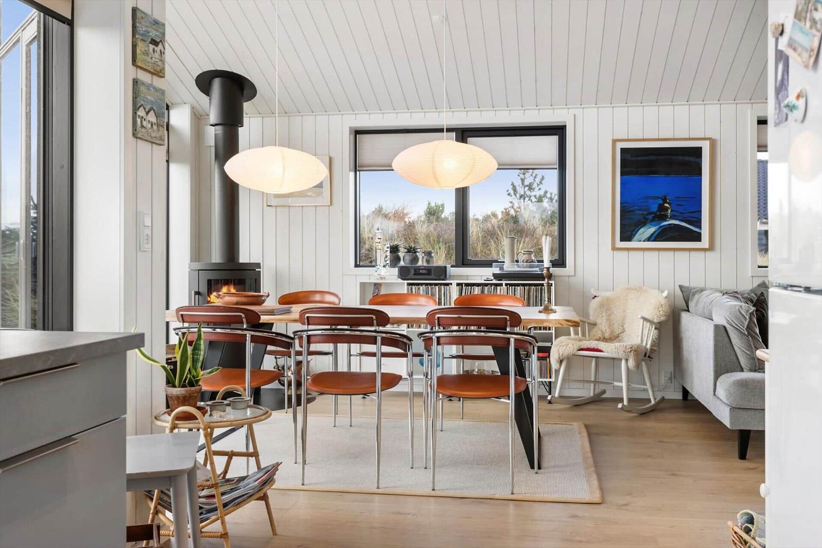 Dining room with wooden table, chairs, and fireplace. Windows overlook green areas.