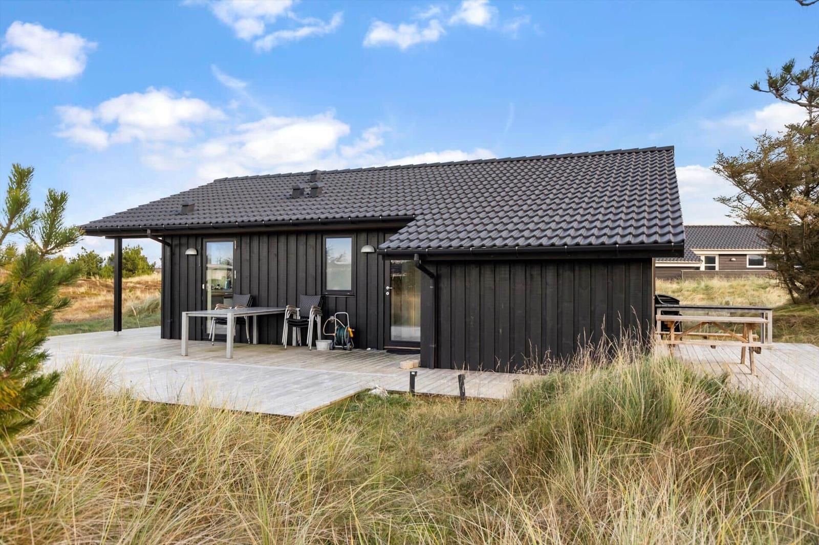 Black house with terrace and garden. Table, chairs, and grill are present.