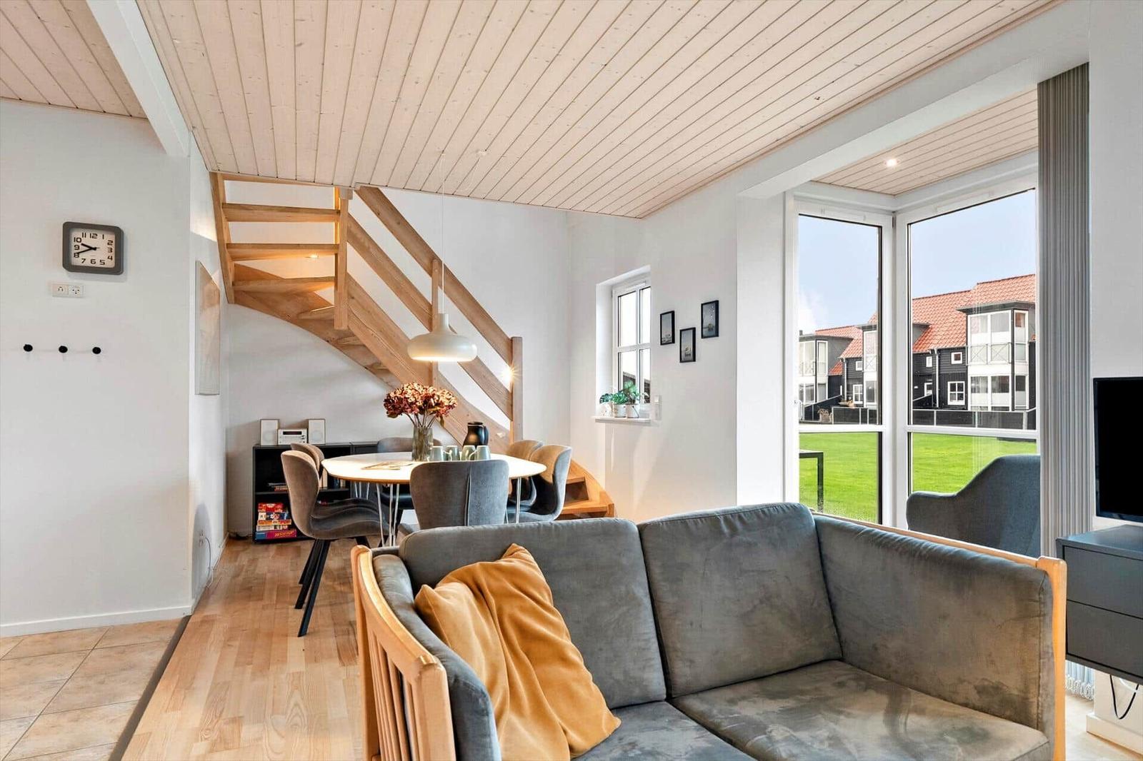 Living and dining area with wooden ceiling, sofa, and table. Large windows show garden and houses.