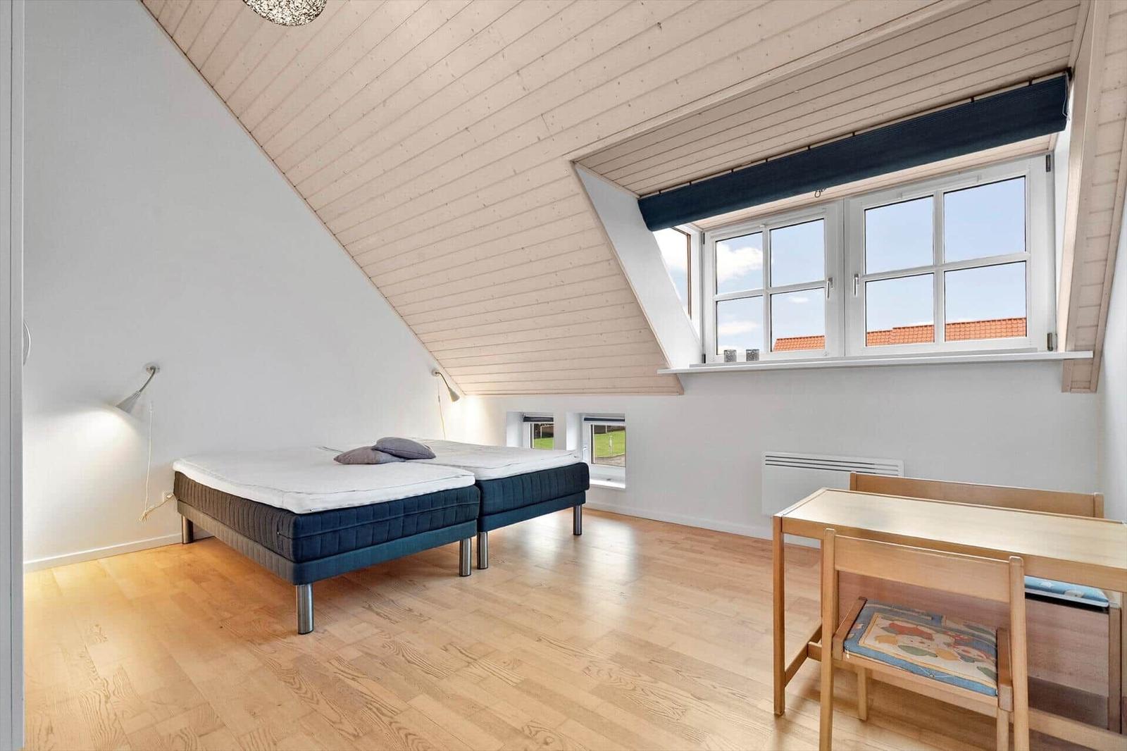 Room with sloped ceiling, double bed, table and chair. Windows overlook rooftops.