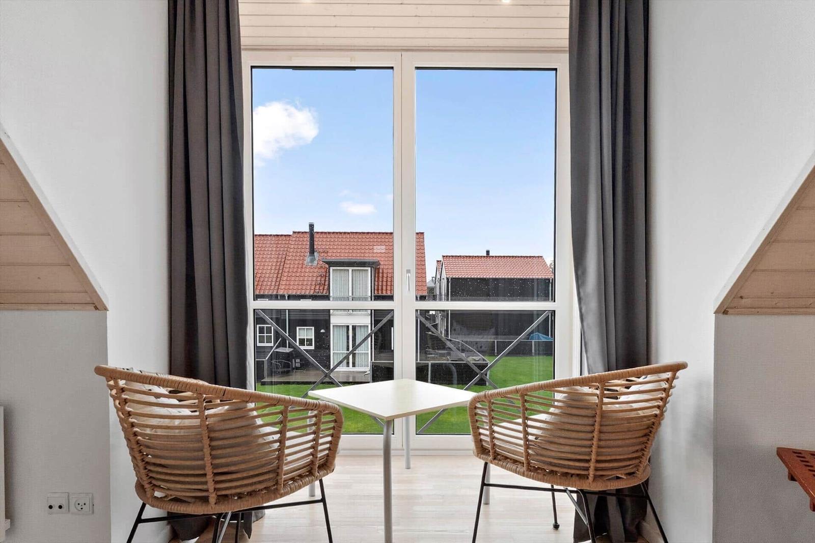 Two rattan chairs and a table in front of a large window with view of red roofs.