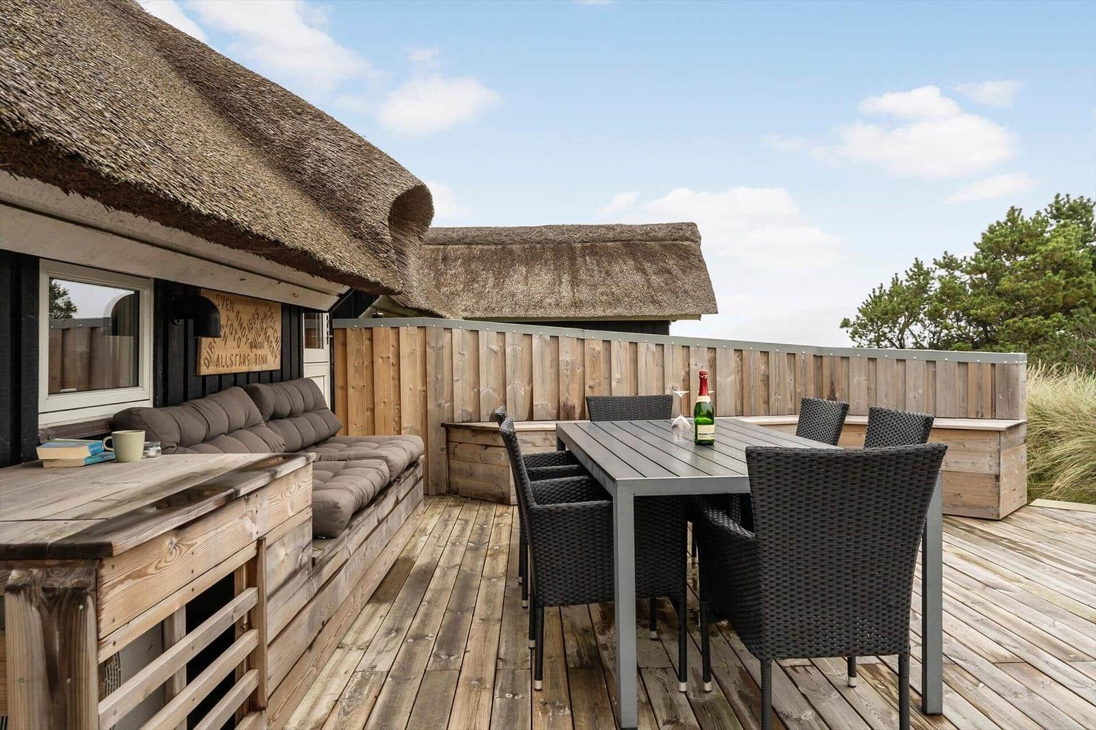 Terrasse mit Sitzbank, Tisch und Stühlen. Hintergrund: Holzzaun und Strohdach.