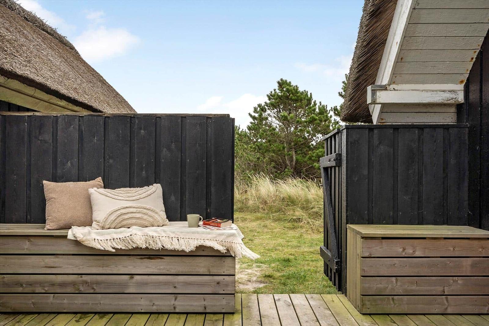 Holzbank mit Kissen und Decke auf Terrasse zwischen zwei Häusern mit Strohdächern.