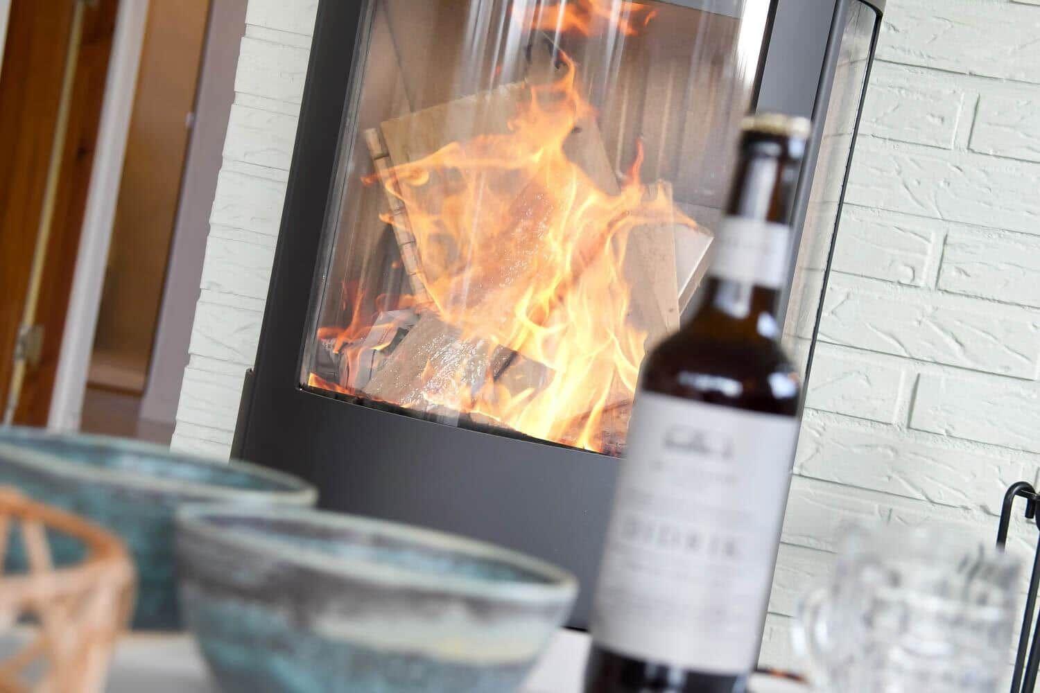 A fireplace with burning logs, a bottle and glasses on a table.