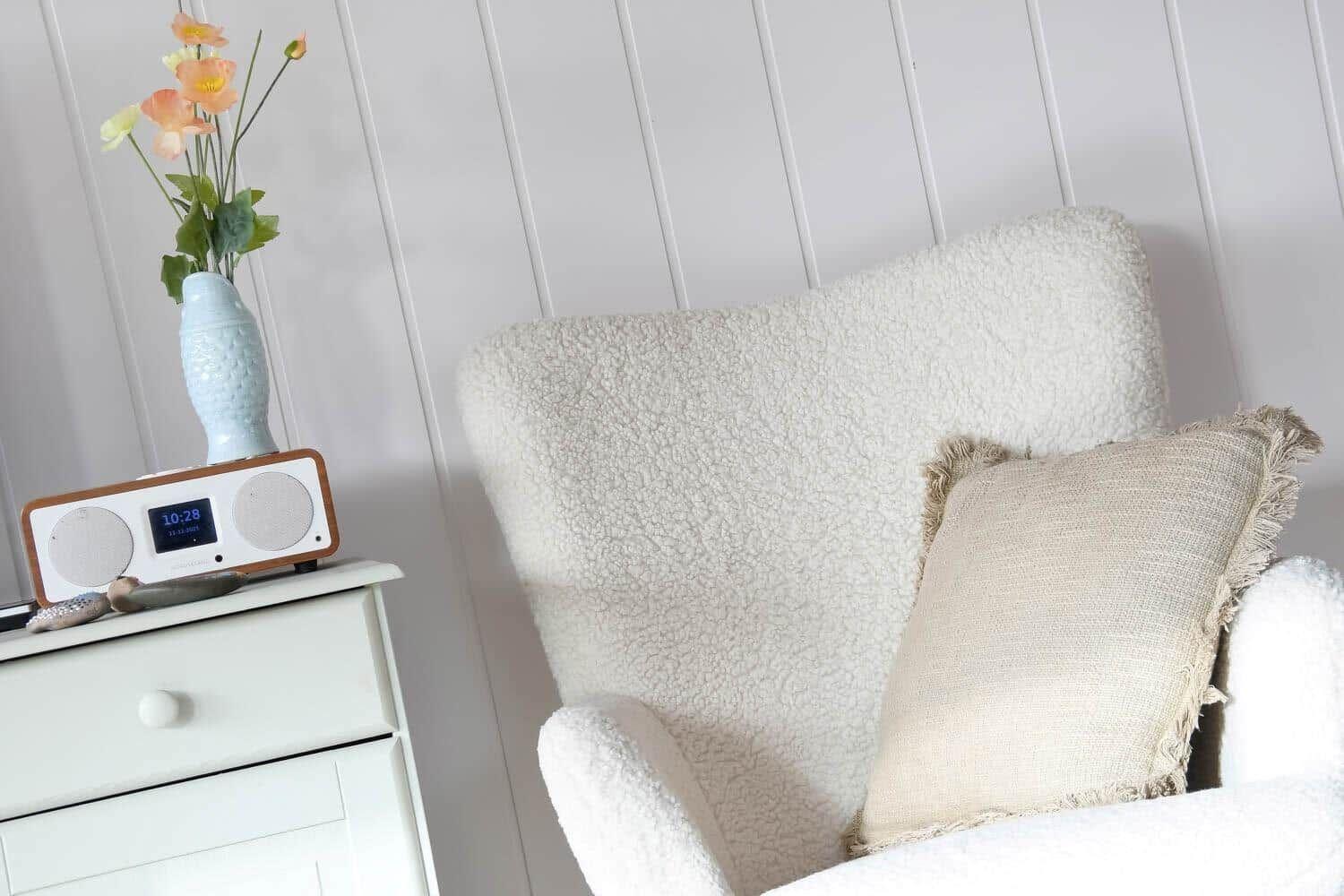 White armchair with cushion, beside nightstand with radio and vase flowers.