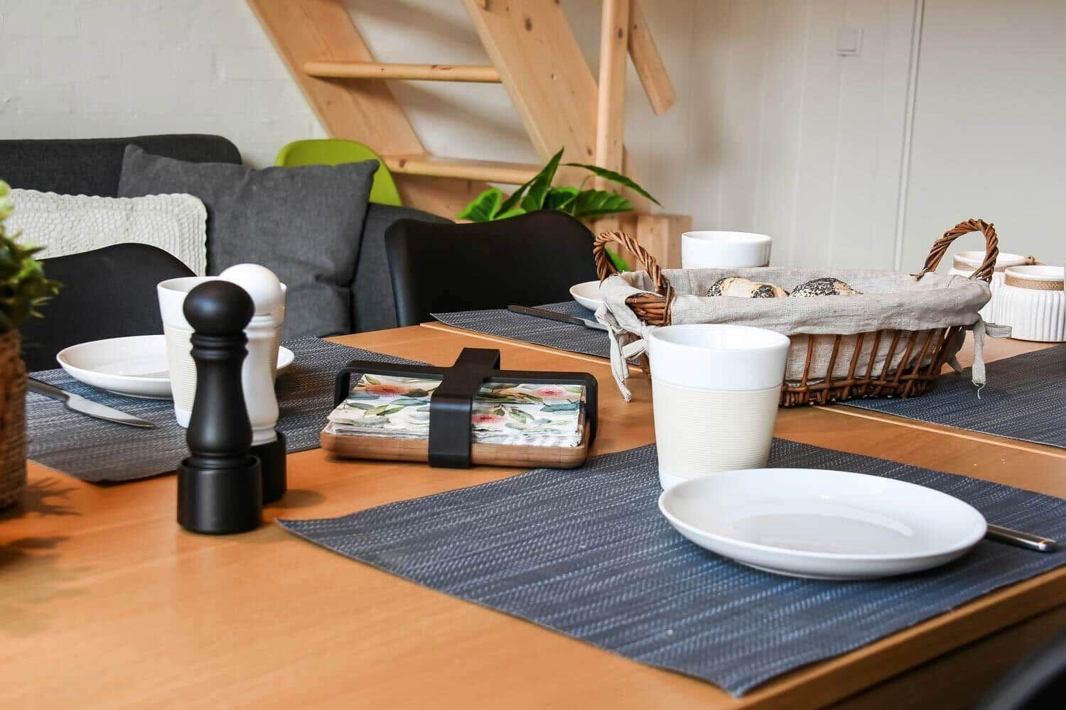 Dining table with placemats, cups, and bread basket. Background: sofa and wooden staircase.