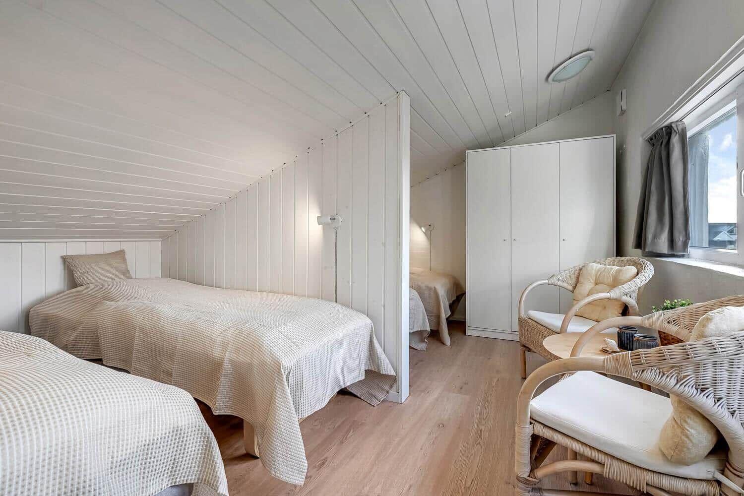 Room with two beds, wardrobe, and seating. Wooden floor and white paneled walls.