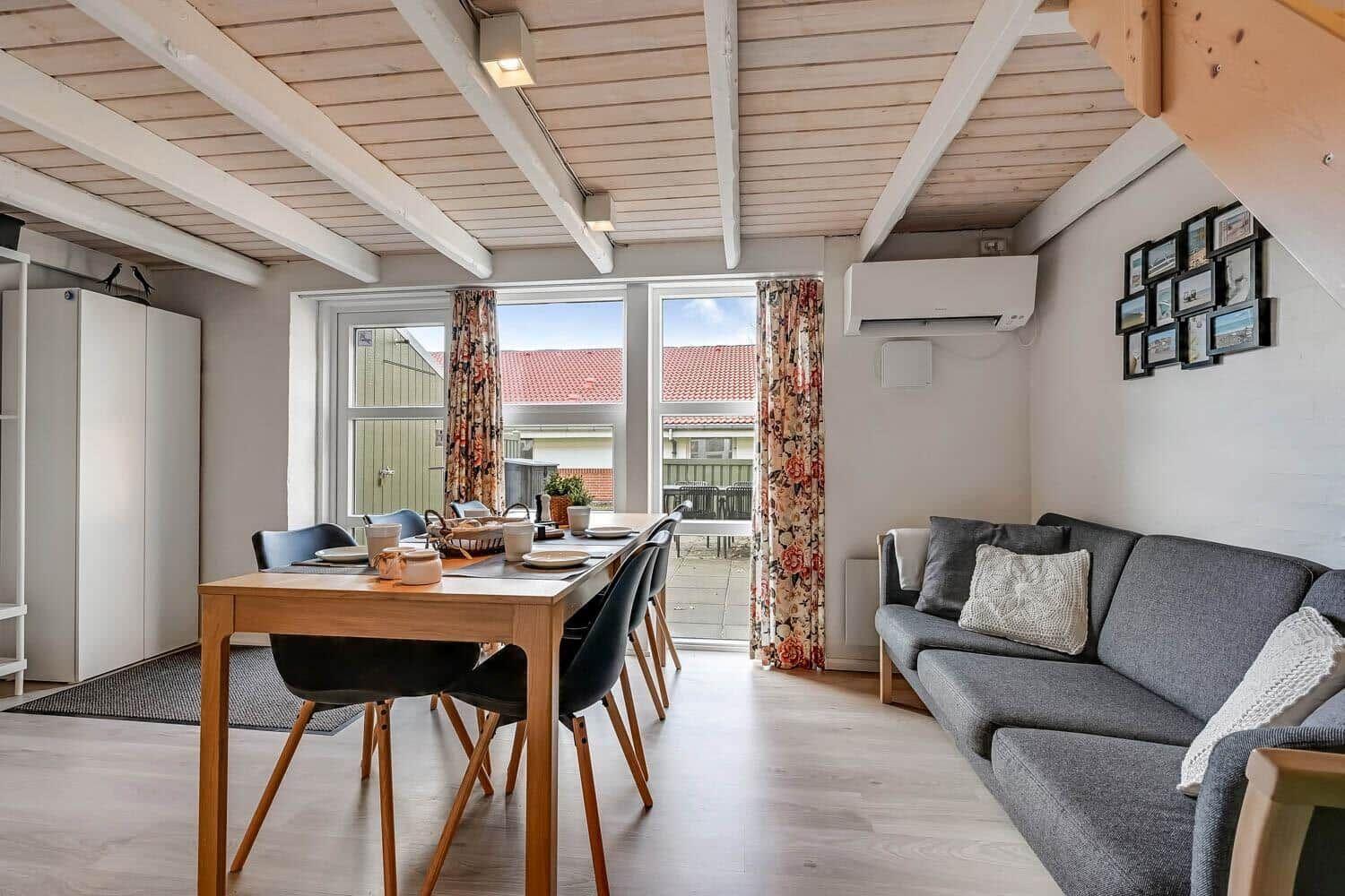 Living and dining area with sofa, table, and windows. Wooden ceiling and floral curtains.