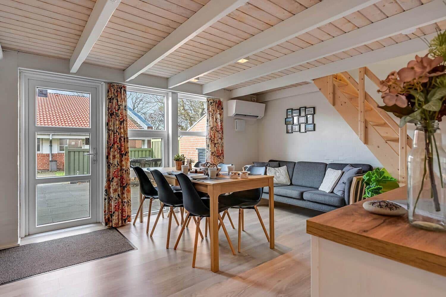 Living and dining area with sofa, table, and chairs. Wooden ceiling and windows with view outside.