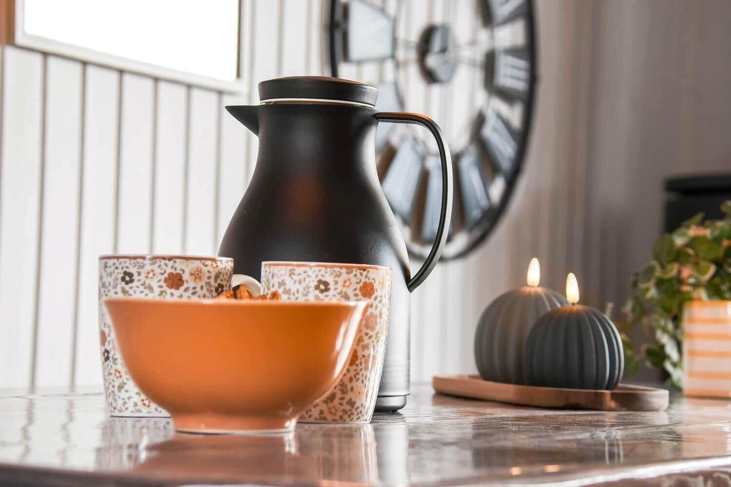 On the table are a mug, bowl, thermos, candles, and plant in the background.