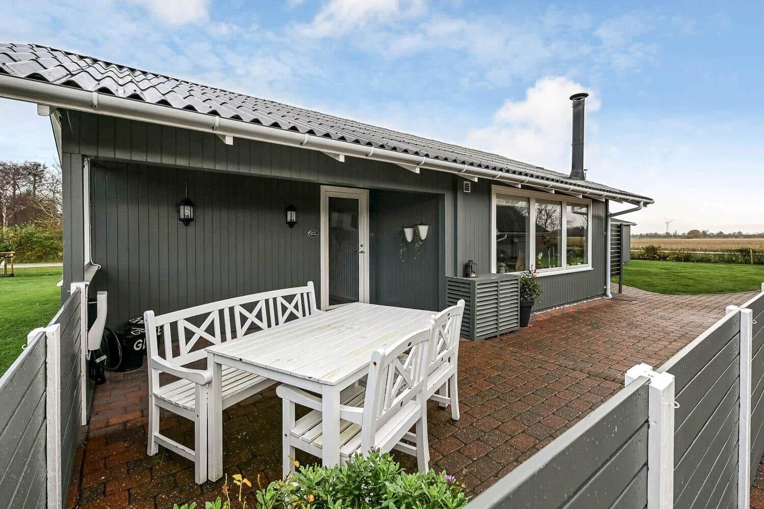 Terrace with white table and chairs in front of gray house with garden.