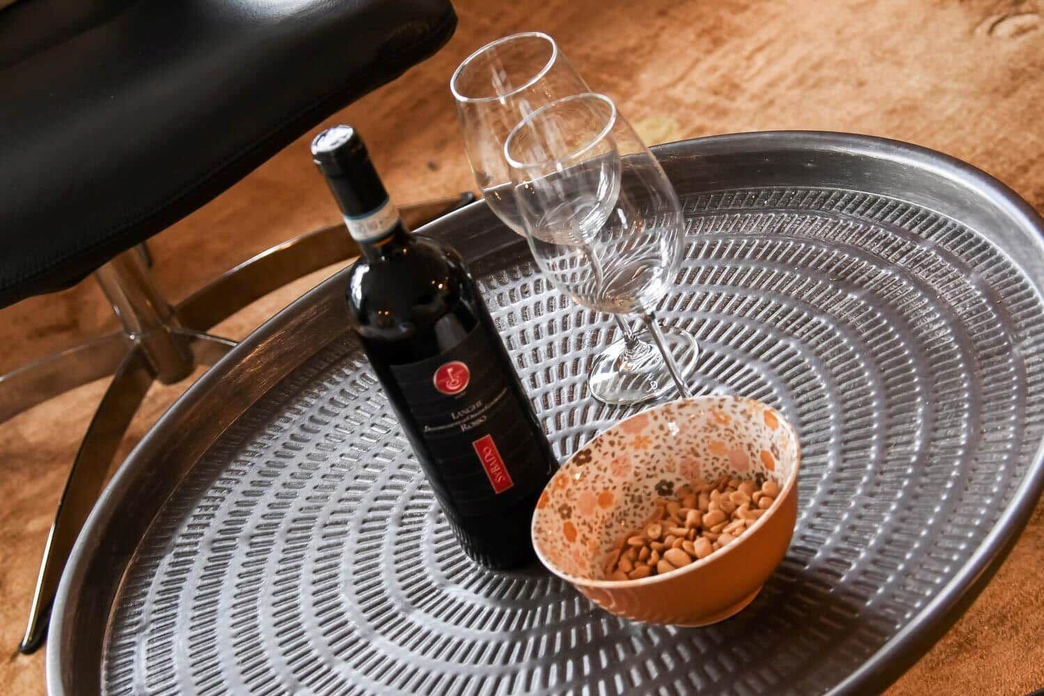 On a metal tray, a wine bottle, two glasses, and a bowl with pine nuts are placed.