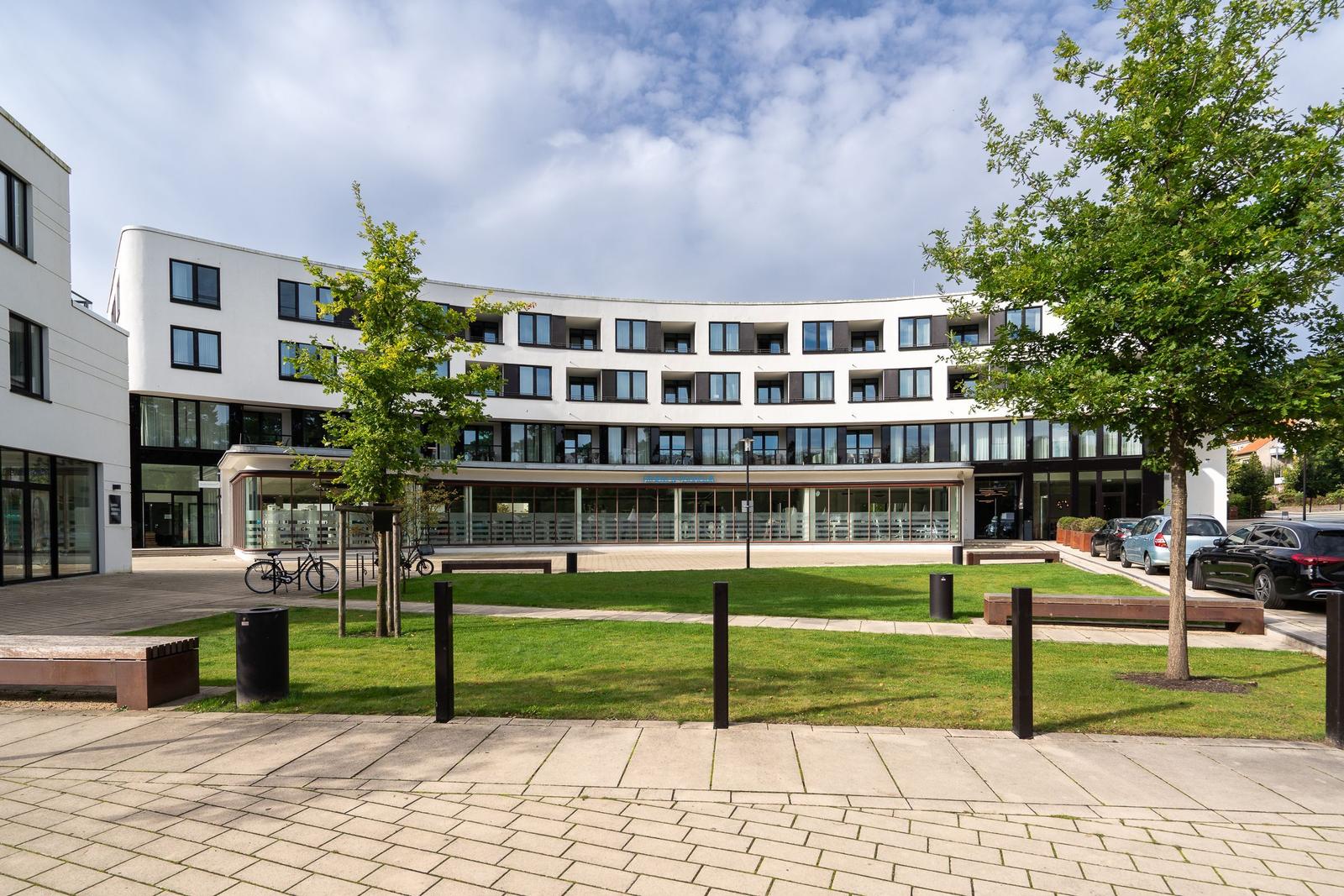 Modernes Gebäude mit Balkonen und Glasfassade. Grünfläche und Parkplätze vorne.