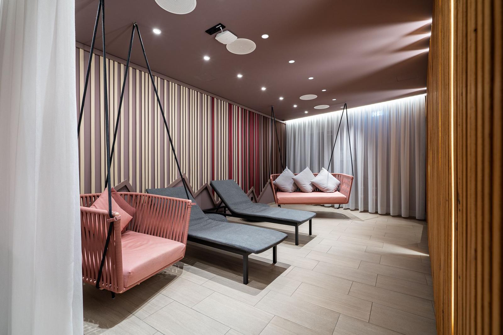 Raum mit hängenden Sitzgelegenheiten und Liegen. Wand mit Streifen, Holzboden und Deckenleuchten.