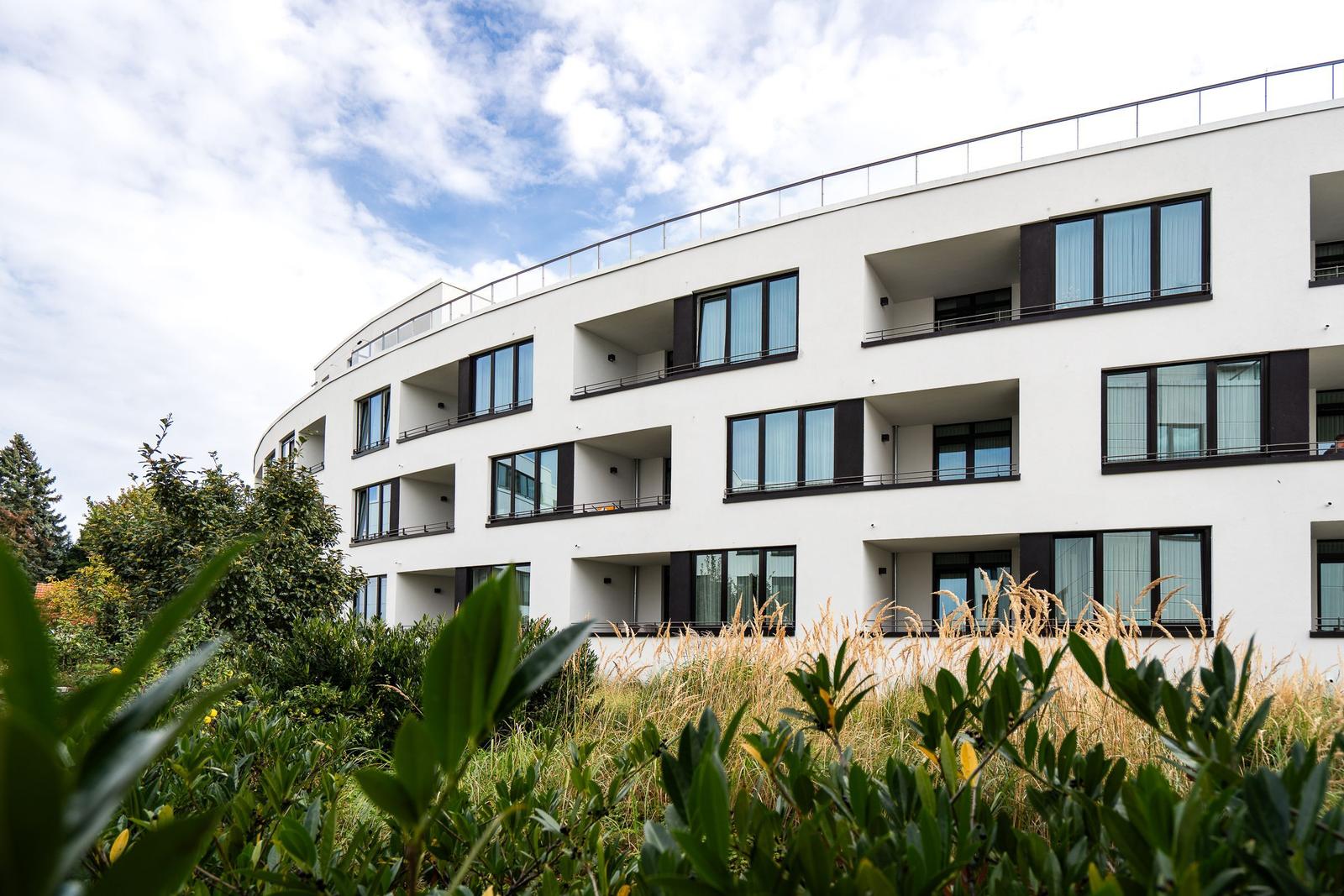 Moderne, weiße Wohnanlage mit Balkonen und großen Fenstern. Grüne Pflanzen im Vordergrund.