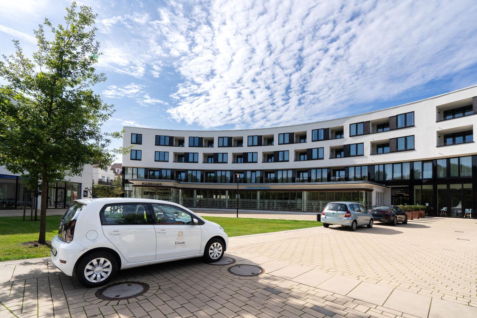 Modernes Gebäude mit gepflastertem Vorplatz und geparkten Autos unter blauem Himmel.