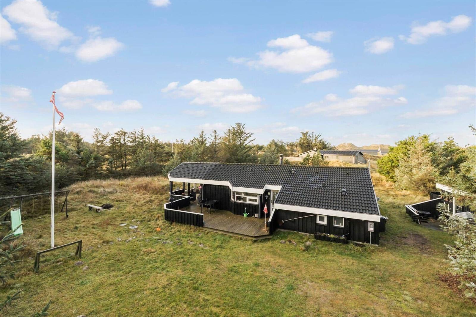 Black house with terrace and garden. A flag stands nearby. Trees and hills are visible in the background.