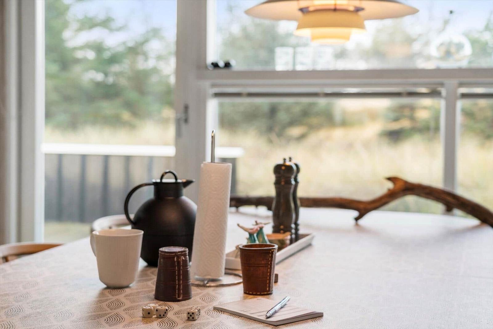 Dining table with tablecloth, coffee pot, notebook, and dice by window.