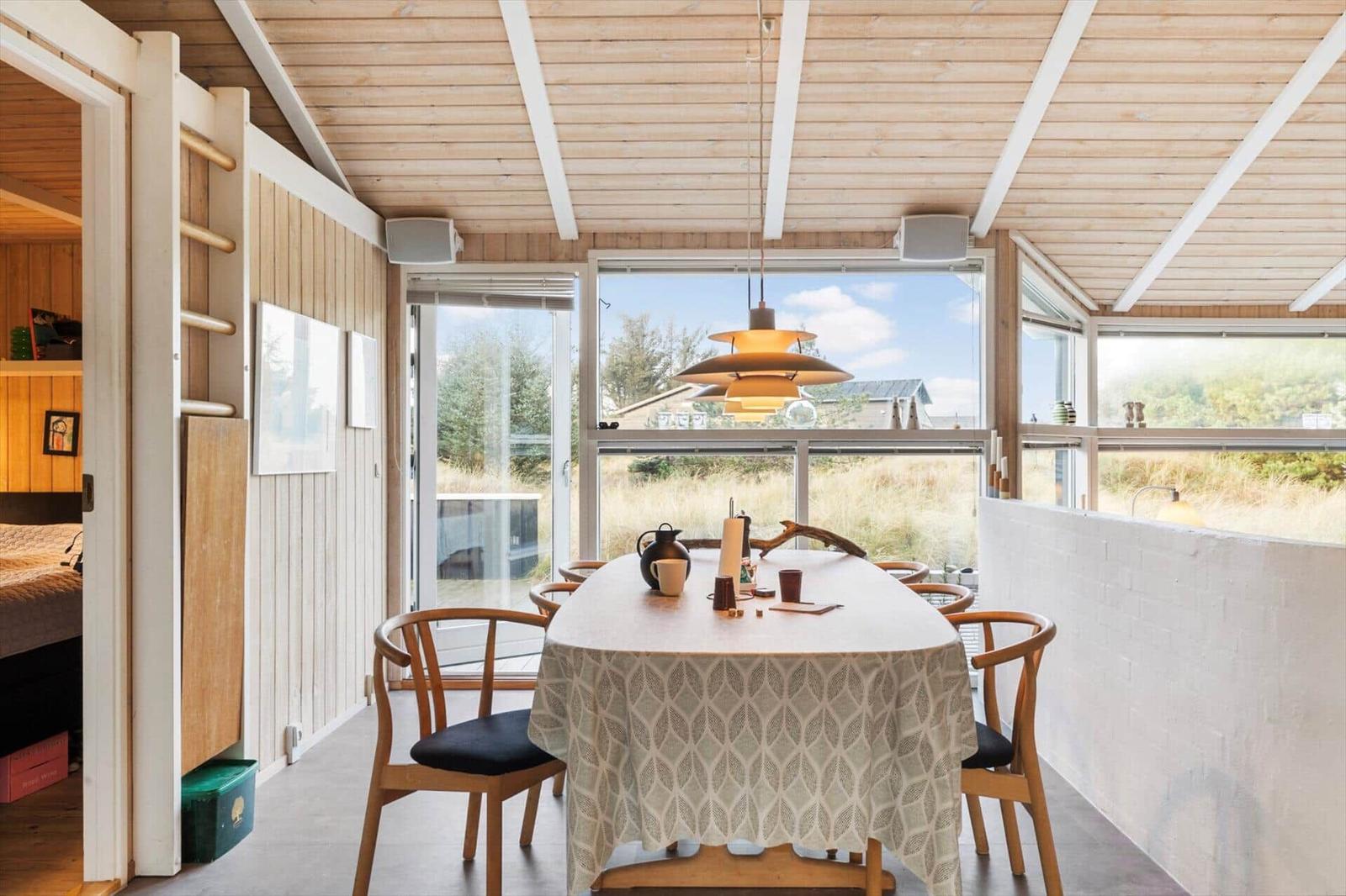 Dining room with table, chairs, and large windows. Wooden ceiling and white wall.
