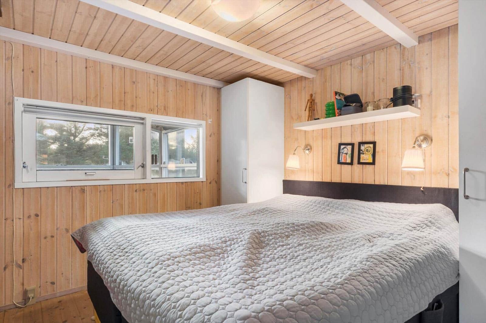 Bedroom with wooden walls, bed, and window. Shelf with items above the headboard.
