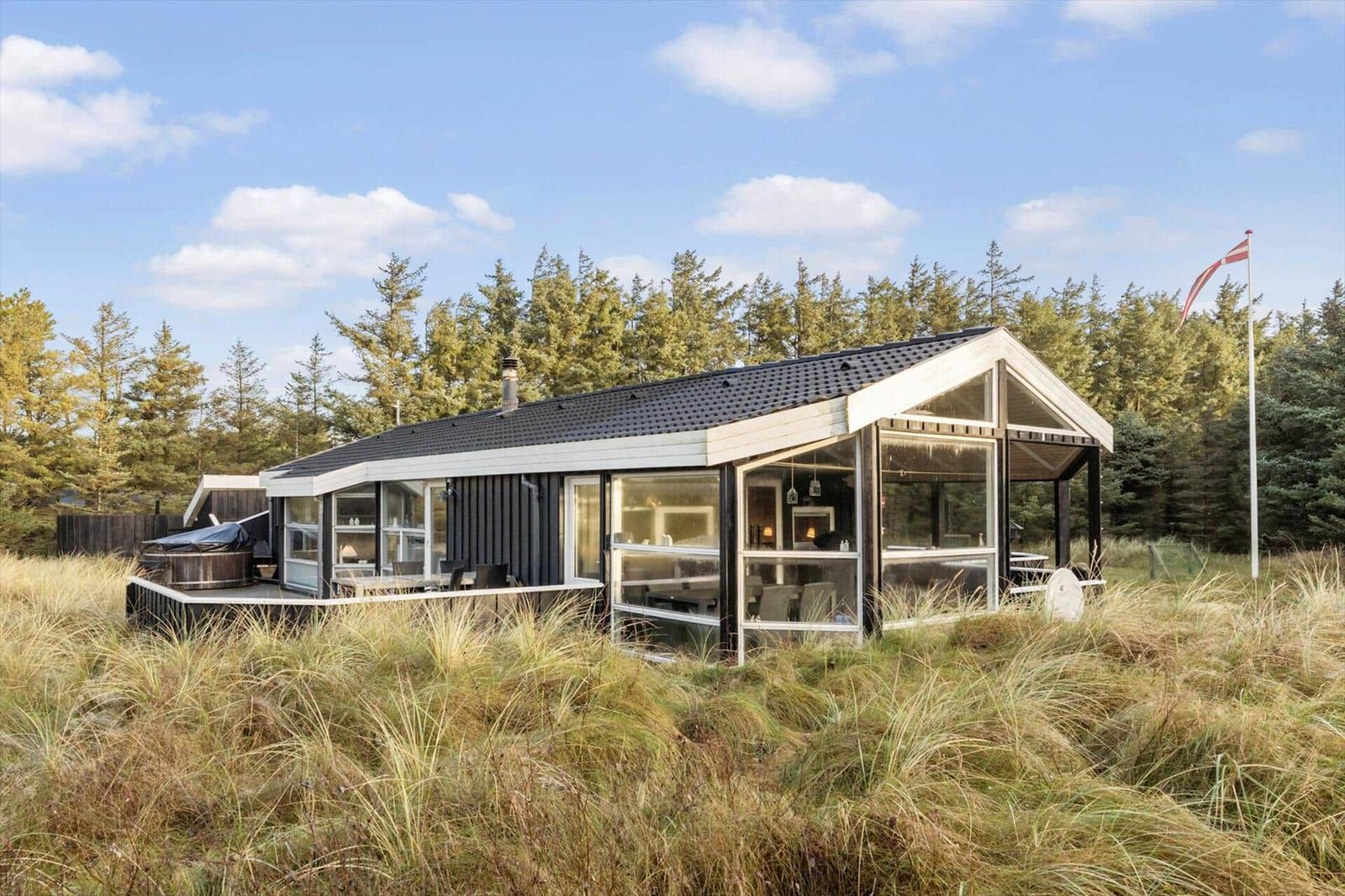 Modern cabin with large glass area, terrace, and hot tub in grassland with forest in background.