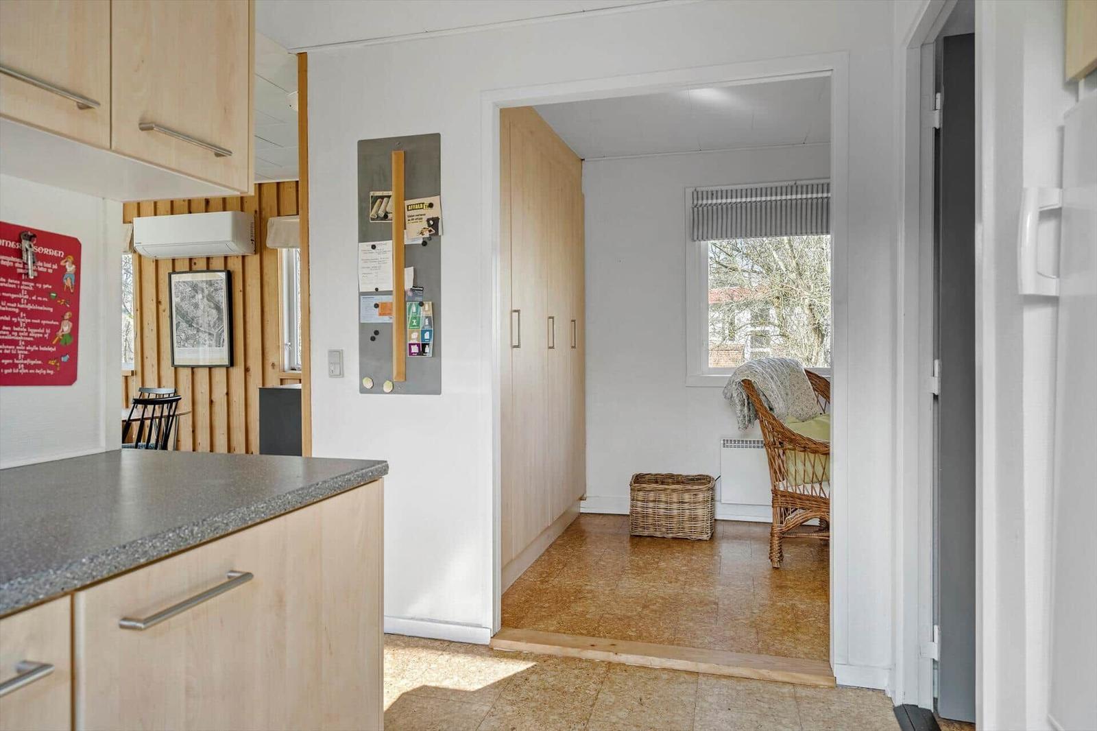 Kitchen with wooden cabinets and view into living area with chair and window.