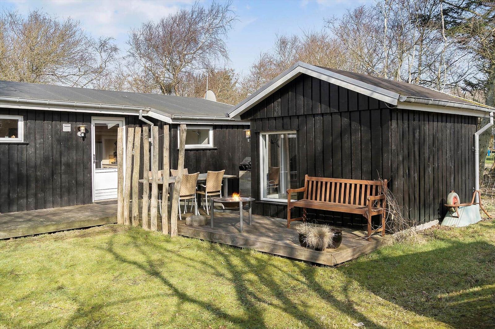 Black wooden house with terrace, bench, and fire pit in the garden.