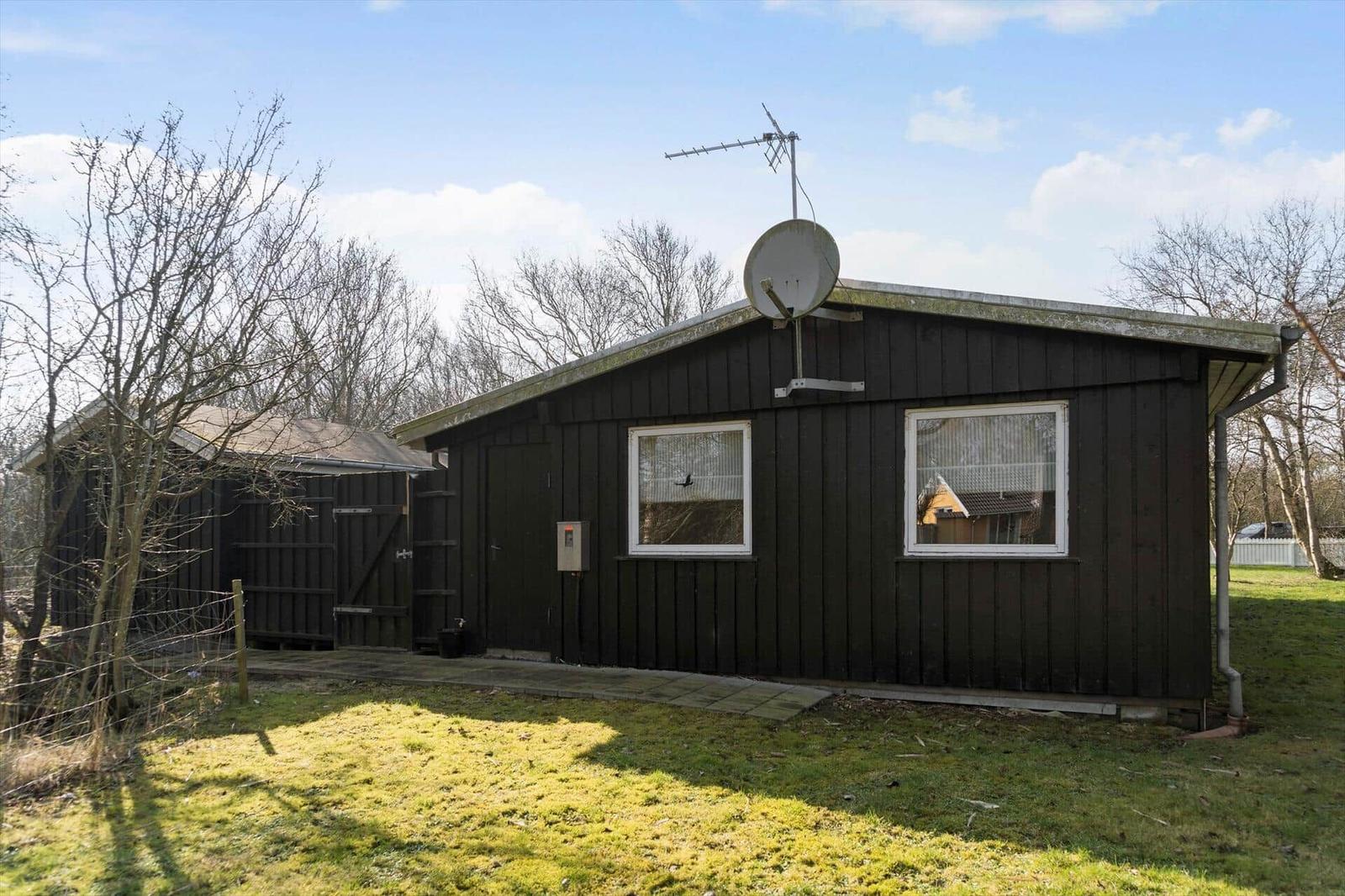 Black wooden house with satellite dish and two windows. Lawn and trees in the background.