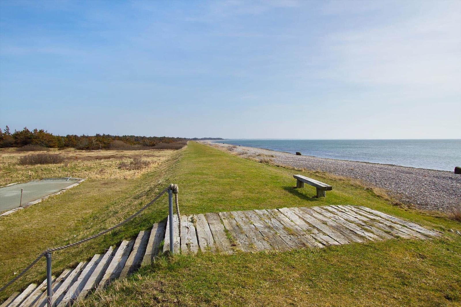 Wooden path leads to the beach. Grass area, bench, and view of the sea.