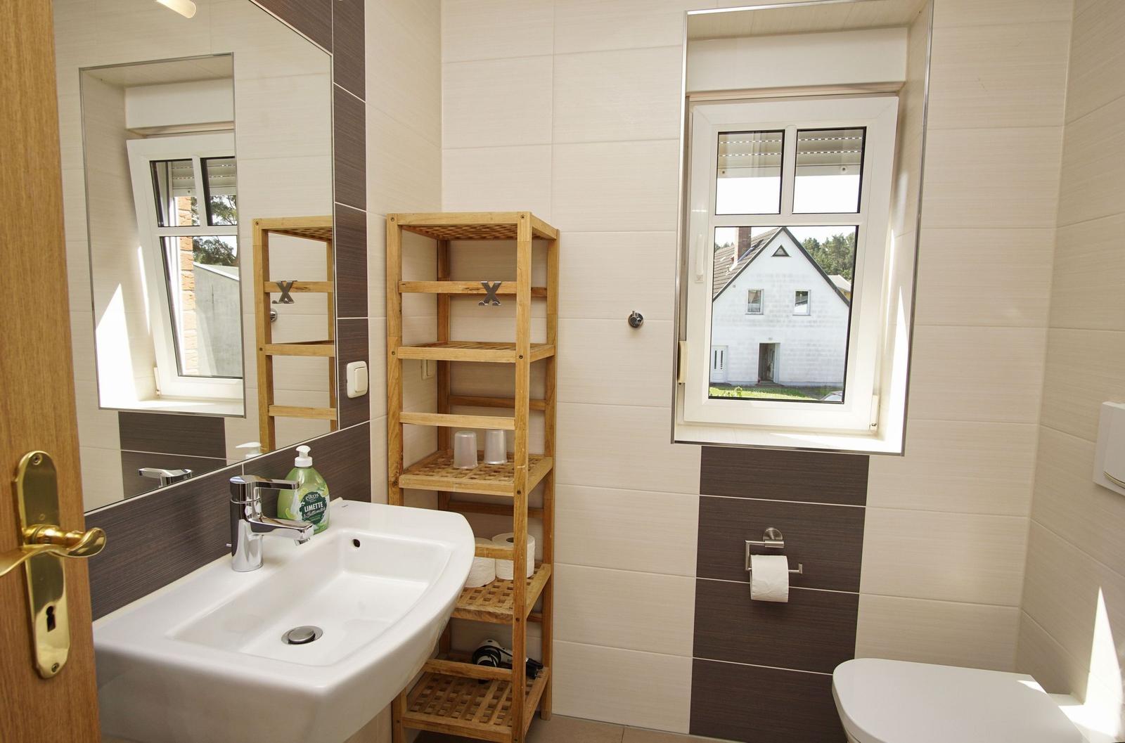 Badezimmer mit Waschbecken, Spiegel, Holzregal und Fenster mit Blick auf Haus.