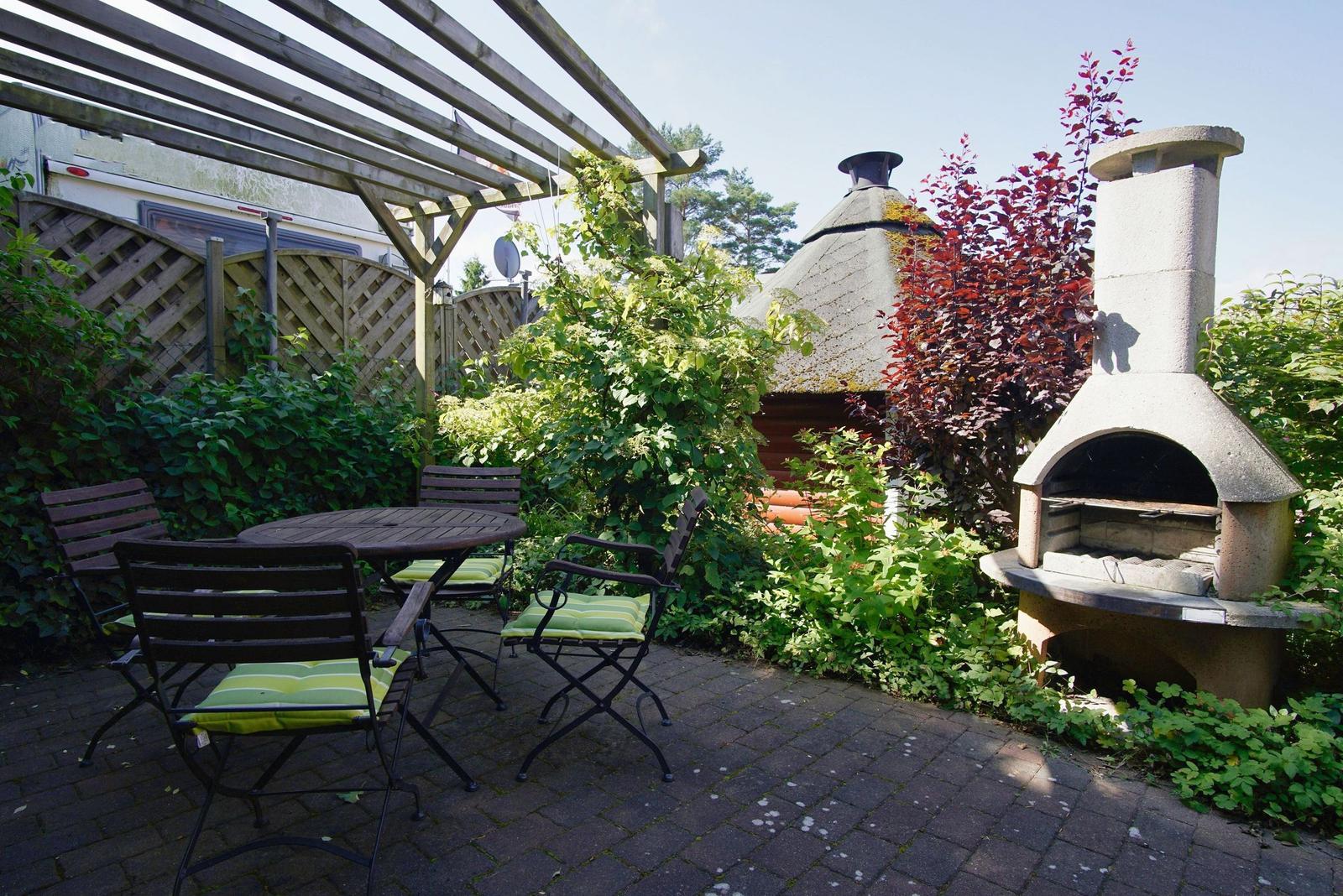 Terrasse mit Tisch, Stühlen und Grill, umgeben von Pflanzen und einem Pavillon.