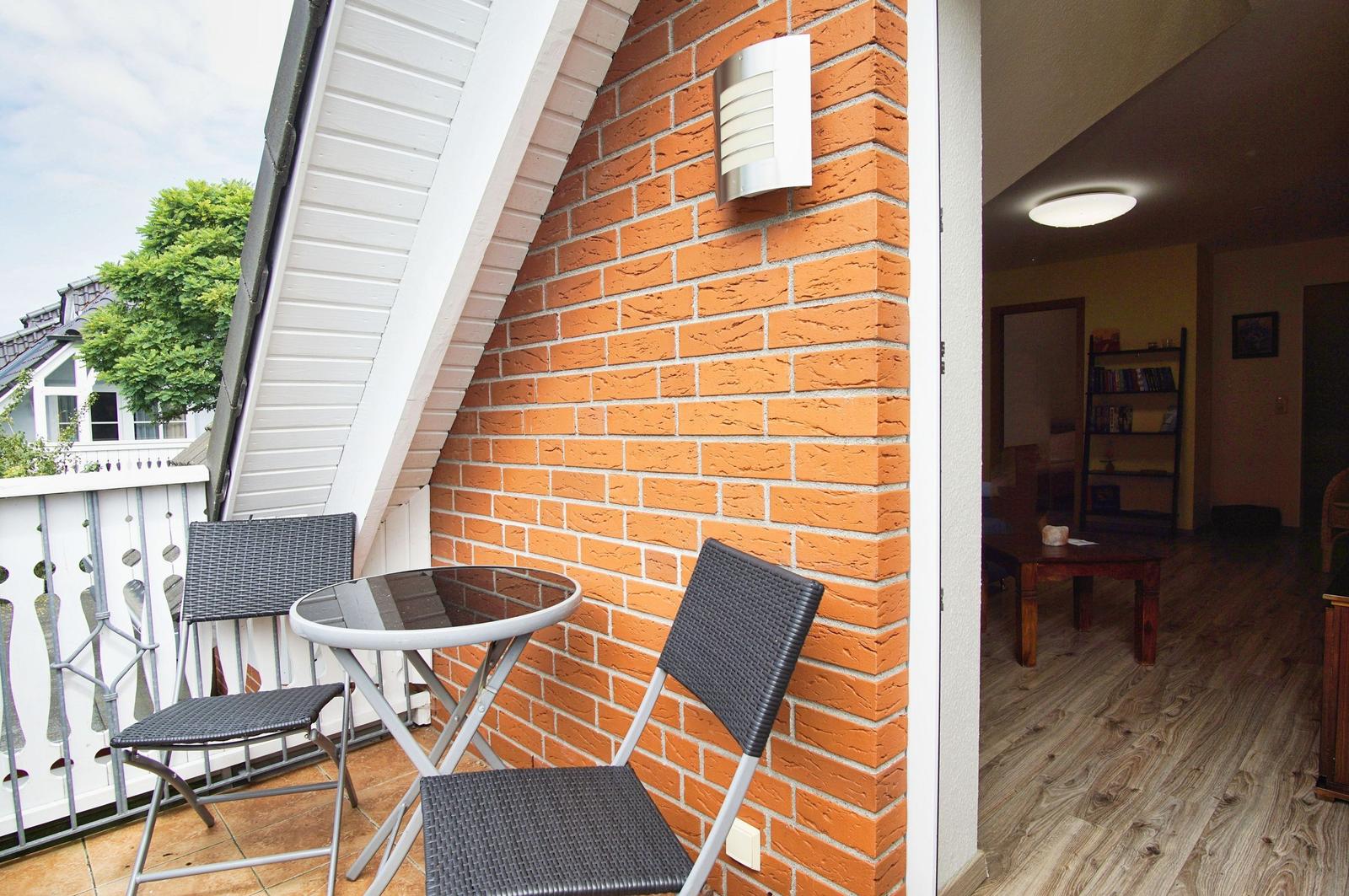 Terrasse mit Tisch und zwei Stühlen neben roter Ziegelwand. Blick ins Innere mit Holzboden.
