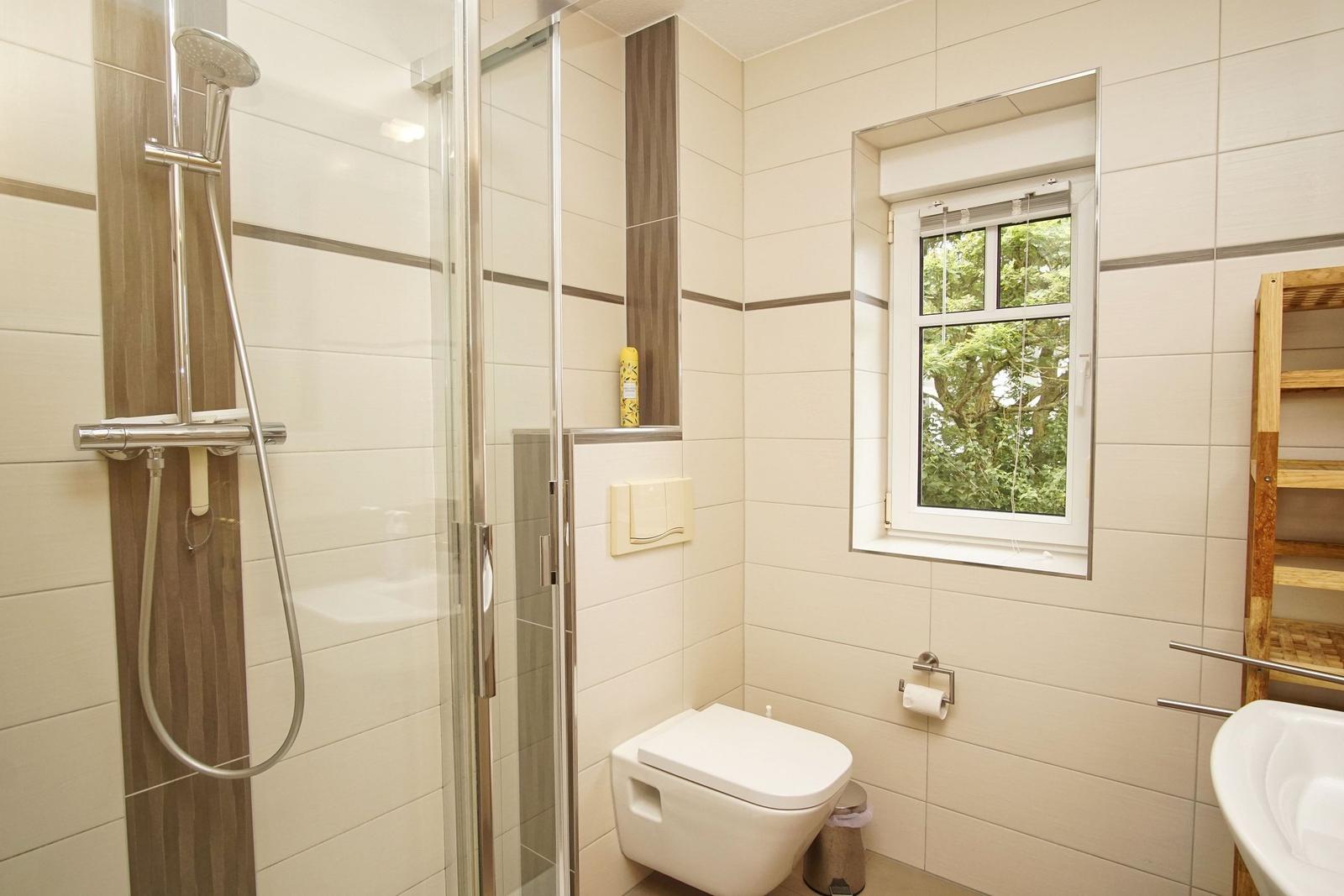 Badezimmer mit Dusche, Toilette und Fenster mit Blick auf Bäume.