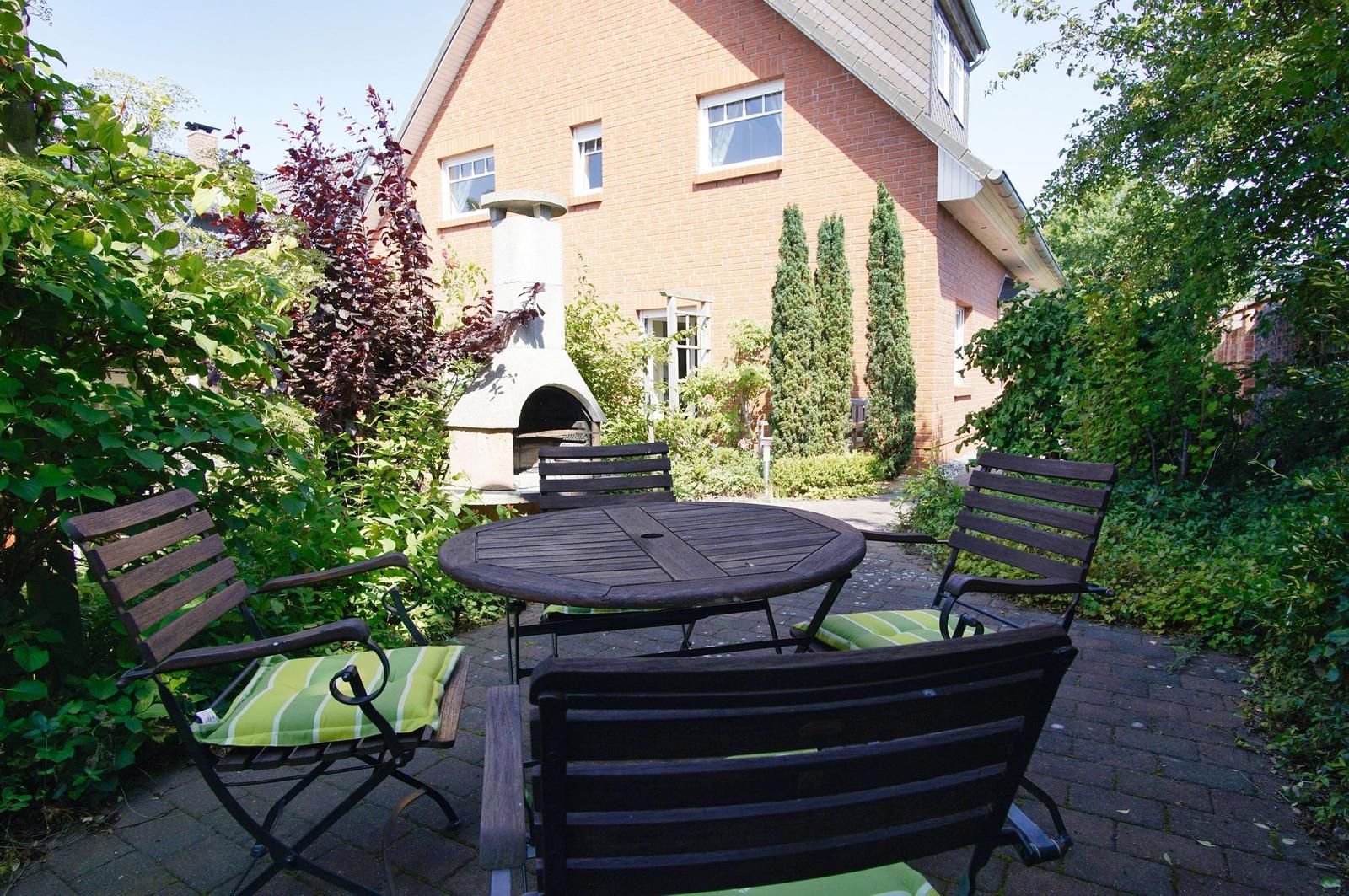 Garten mit Holztisch, Stühlen und Grill. Umgeben von Bäumen und Sträuchern.