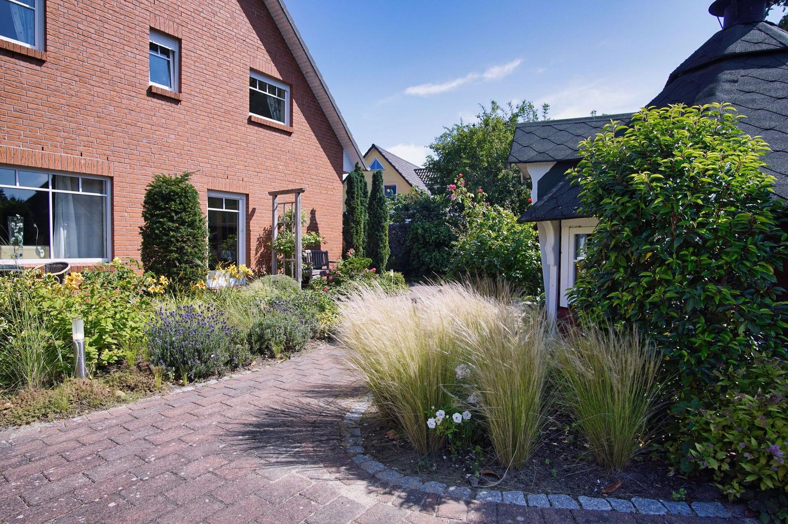 Pflasterweg durch einen Garten mit Blumen und Gräsern vor einem roten Backsteinhaus.