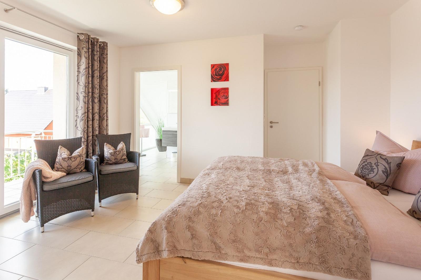 Schlafzimmer mit Bett, zwei Sesseln und Blick auf Balkon.