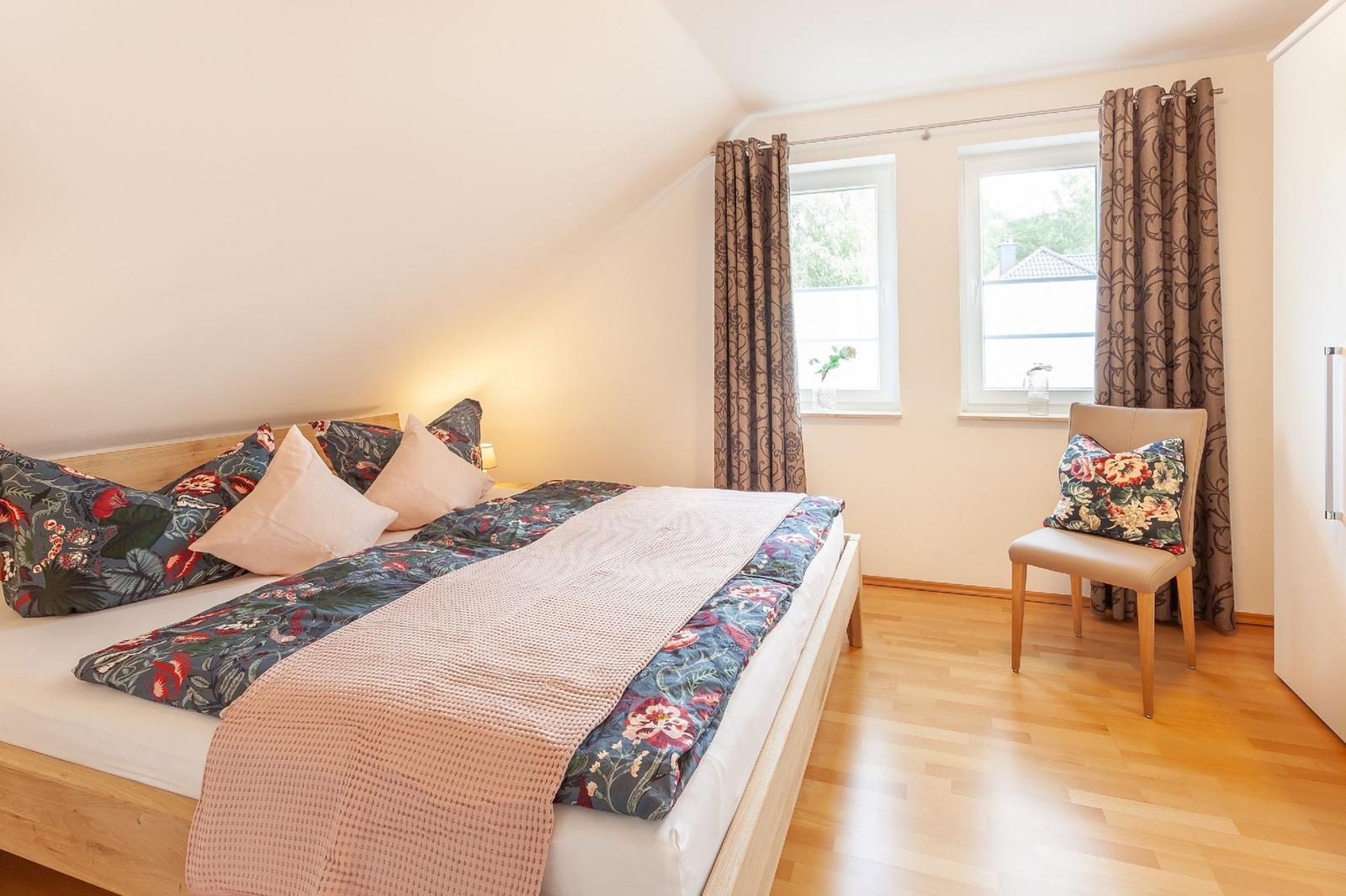 Schlafzimmer mit Bett, Stuhl und zwei Fenstern mit Vorhängen.