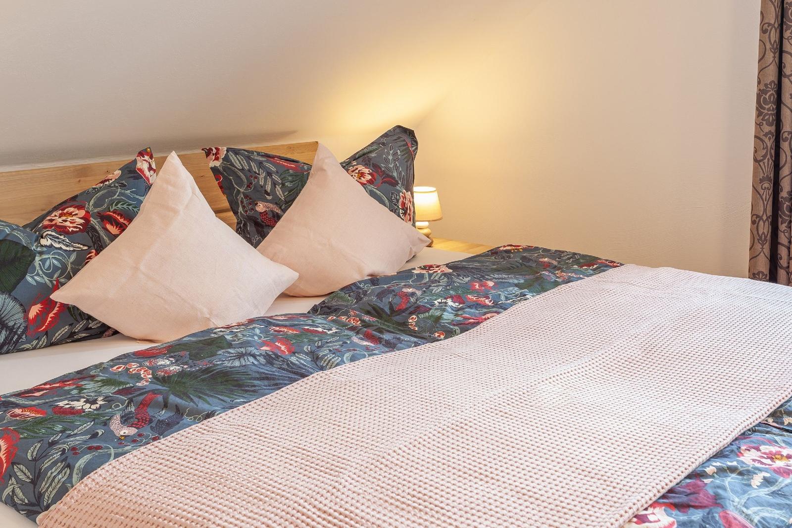 Das Bett ist mit einem floralen Bettbezug und Kissen bezogen. Eine Nachttischlampe steht daneben.