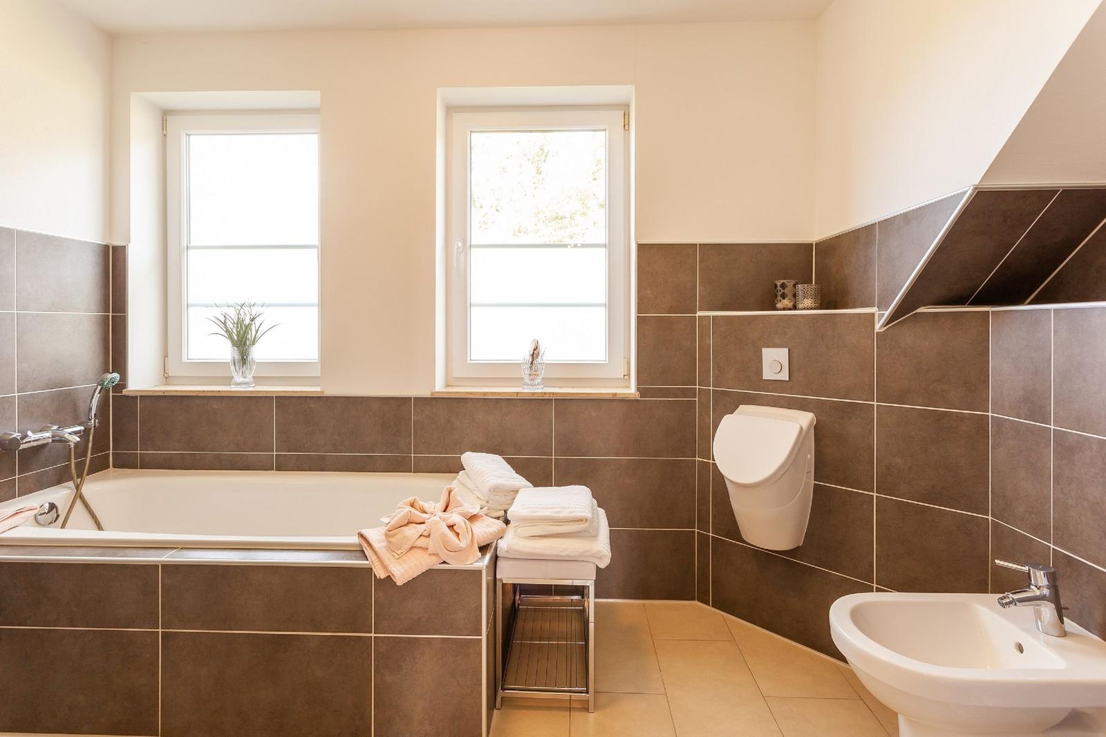 Badezimmer mit Badewanne, Toilette und Waschbecken. Zwei Fenster und braune Fliesen.