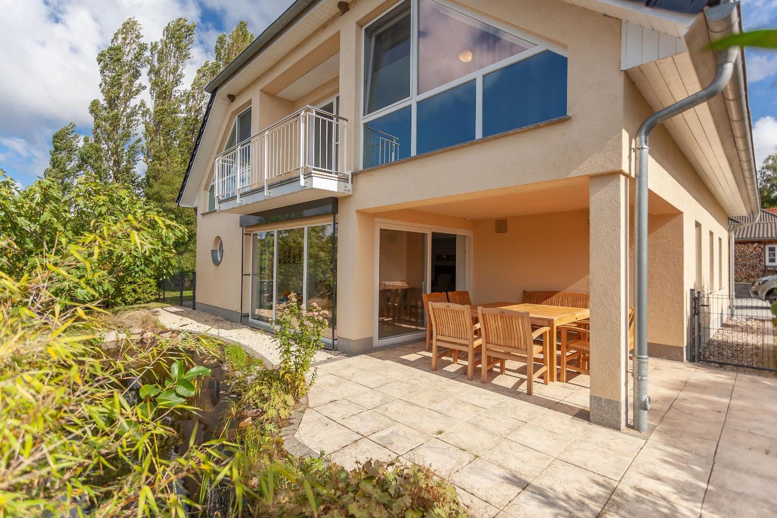 Haus mit Terrasse, Tisch und Stühlen, kleinem Teich und Balkon im Hintergrund.
