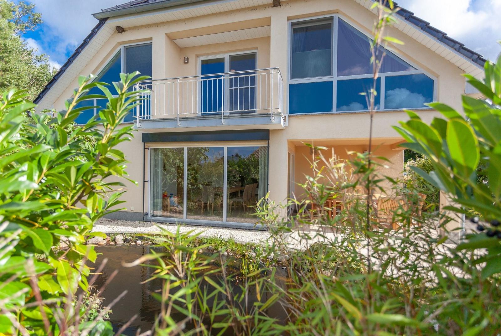 Modernes Haus mit Balkon und Terrasse, umgeben von Grün und einem kleinen Teich.