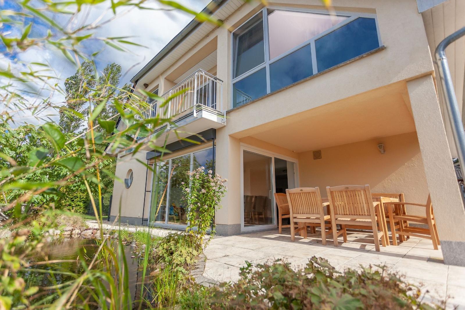 Haus mit Terrasse, Garten und Holzmöbel. Blick auf Pool und Bäume.