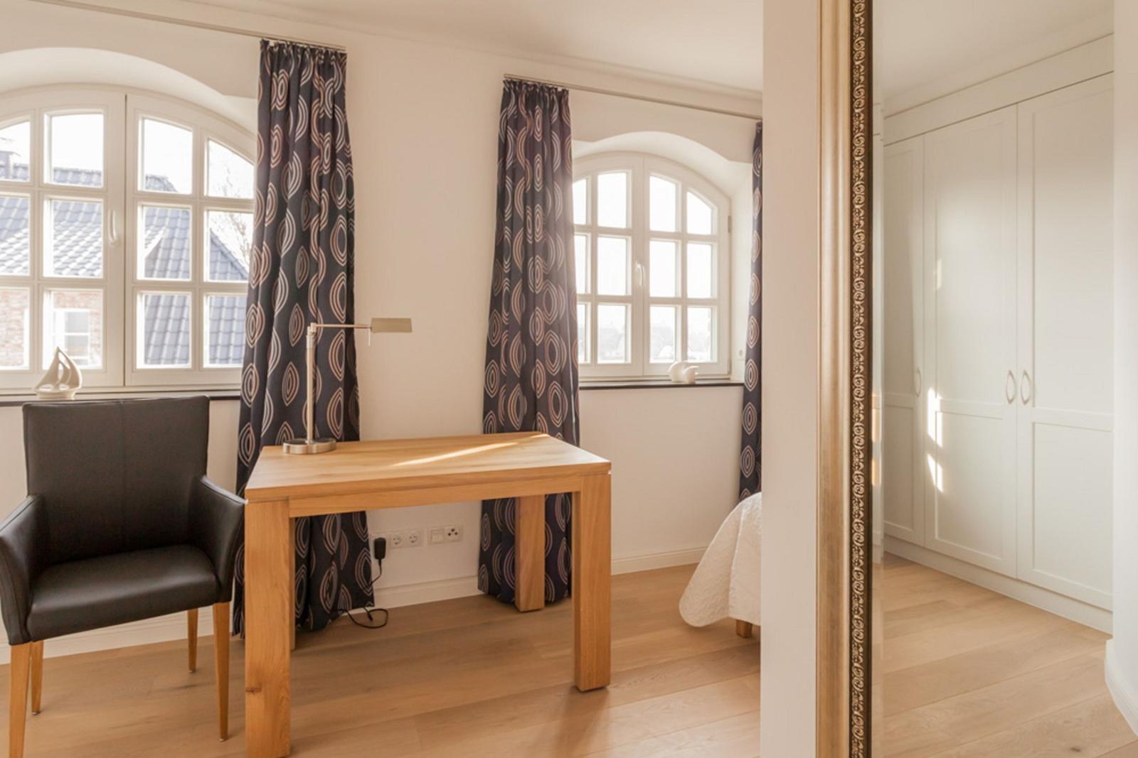 Ein Zimmer mit Schreibtisch, Stuhl und Fenstern. Spiegel zeigt Kleiderschrank.