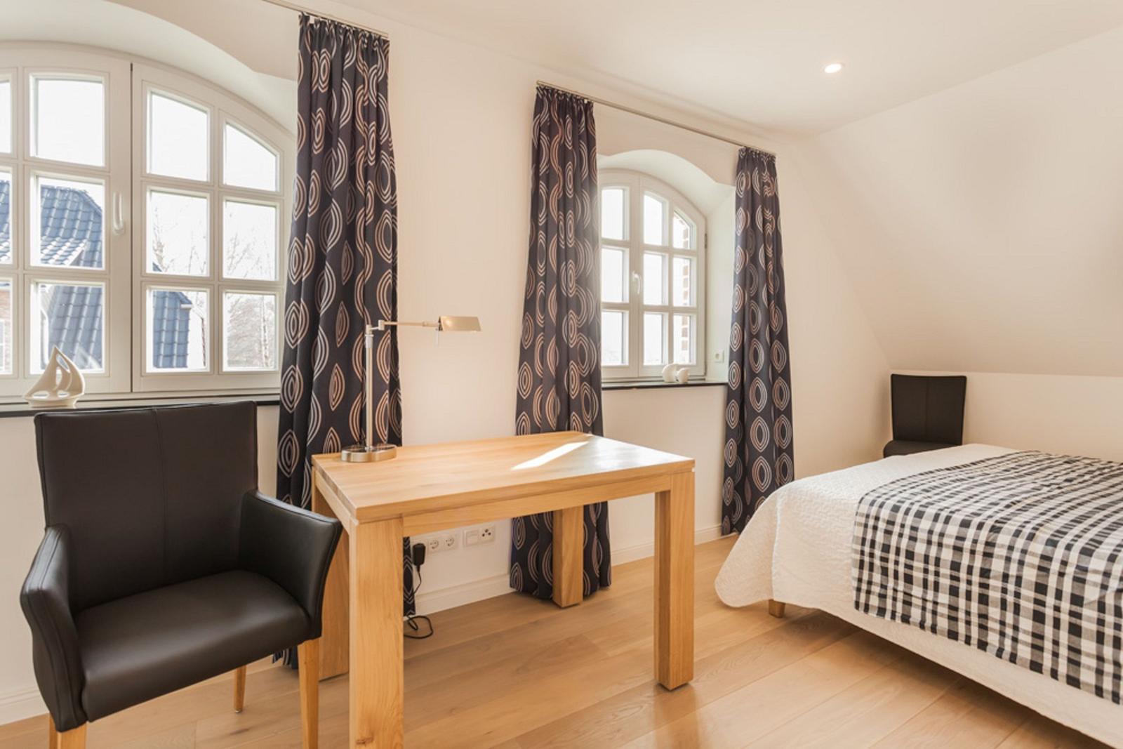 Schlafzimmer mit Bett, Schreibtisch und zwei Fenstern mit Vorhängen.