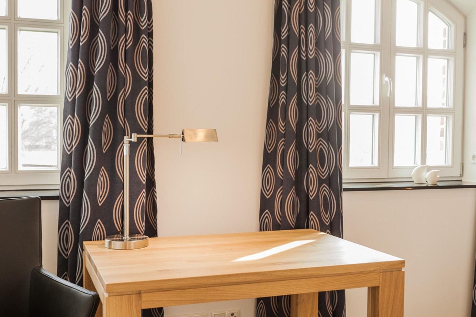 Holztisch mit Lampe zwischen zwei Fenstern mit Vorhängen.