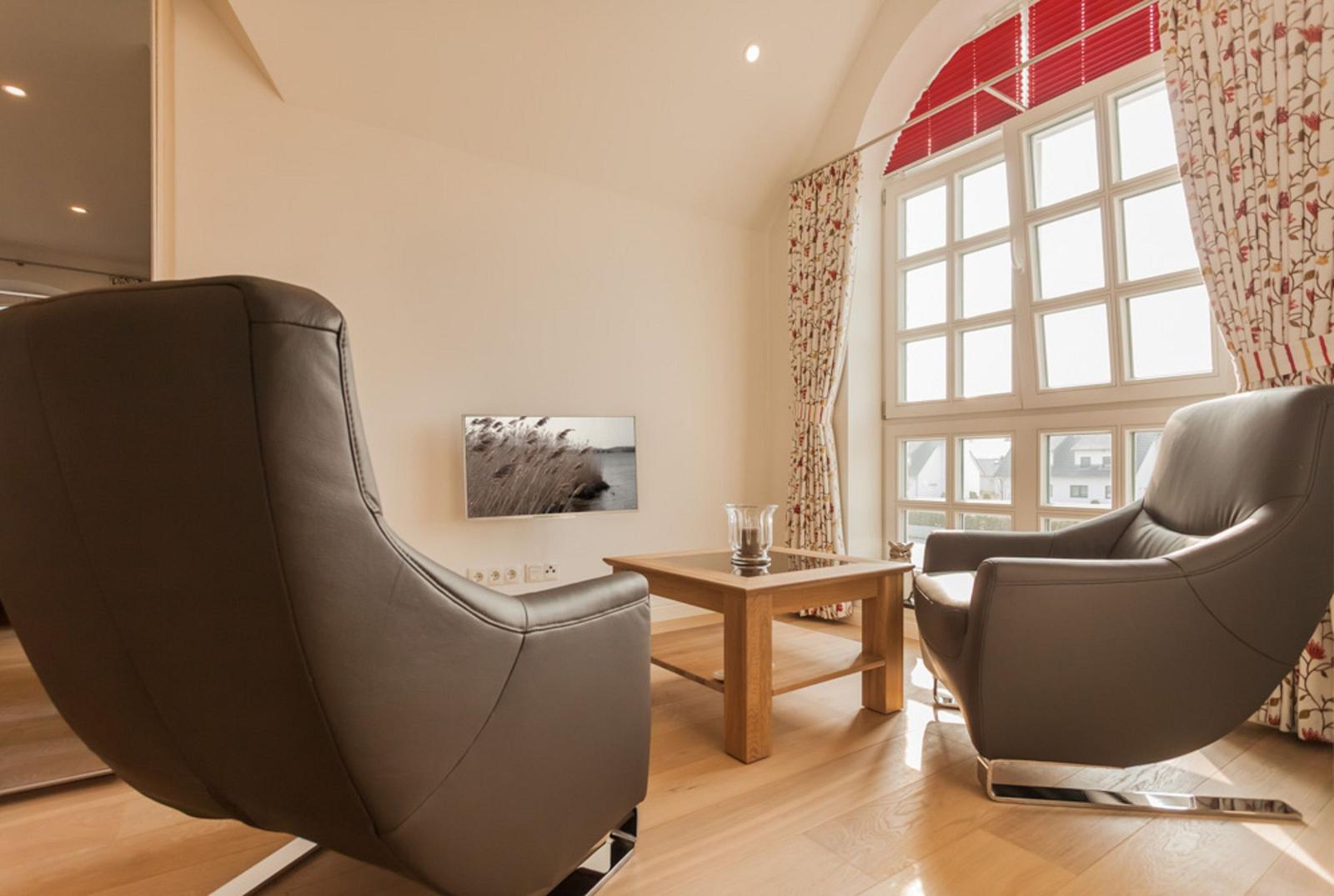 Zwei graue Sessel und ein Holztisch stehen vor einem großen Fenster mit roten Jalousien.